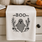 An off-white kitchen towel with a black embroidered 'BOO' design featuring a ghost and leafy wreath, hanging on a wooden kitchen counter with seasonal props like pumpkins and a plant.