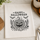 A white kitchen tea towel with a black embroidered 'Happy Halloween' design featuring a smiling jack-o'-lantern, bats, and floral scrollwork, styled on a marble surface with seasonal props.