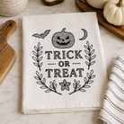 A folded white kitchen towel with black embroidered 'Trick or Treat' text, a jack-o'-lantern, bats, leaves, and a star, styled on a marble countertop with seasonal props.
