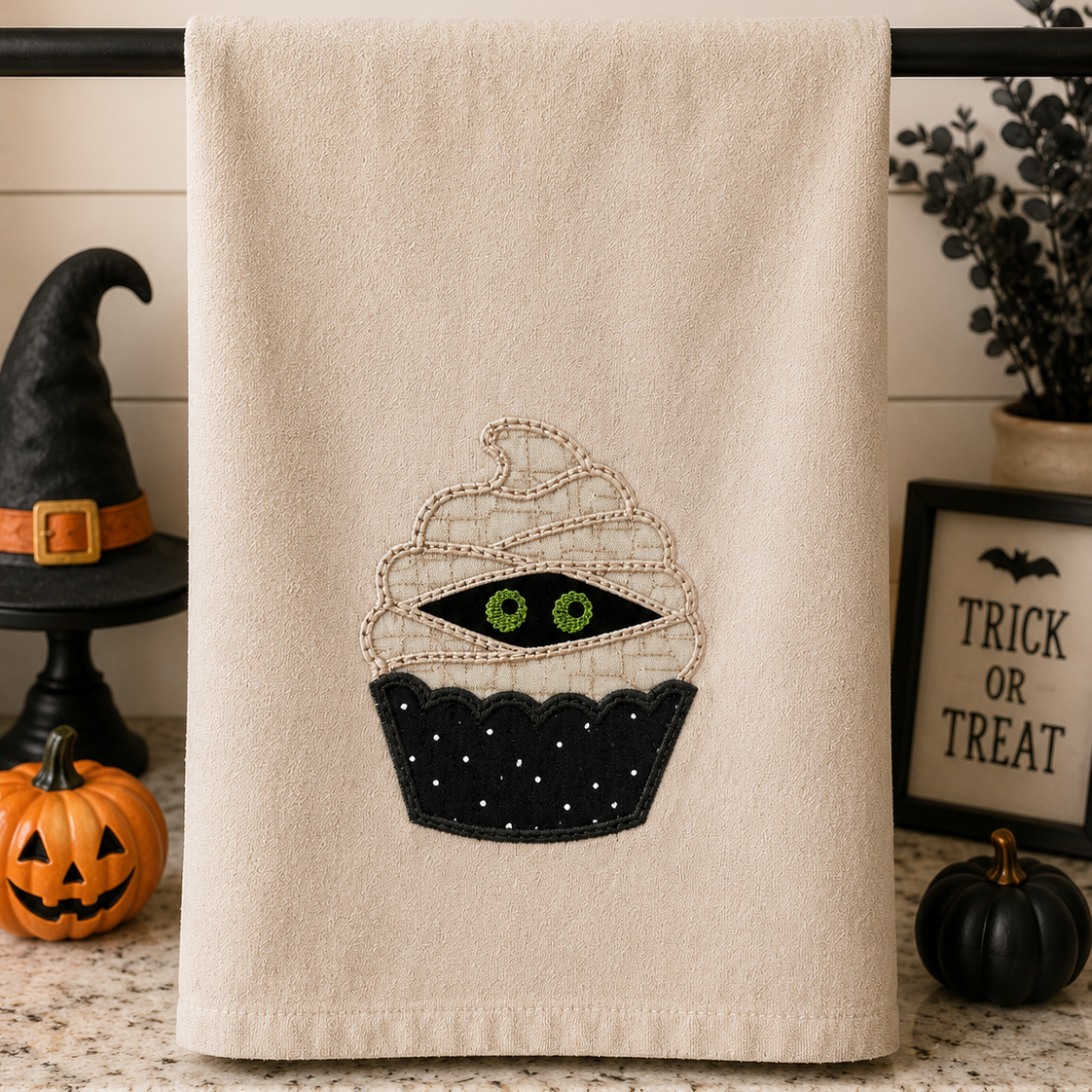 Beige towel with a Halloween-themed cupcake design on a kitchen counter with decorative items.
