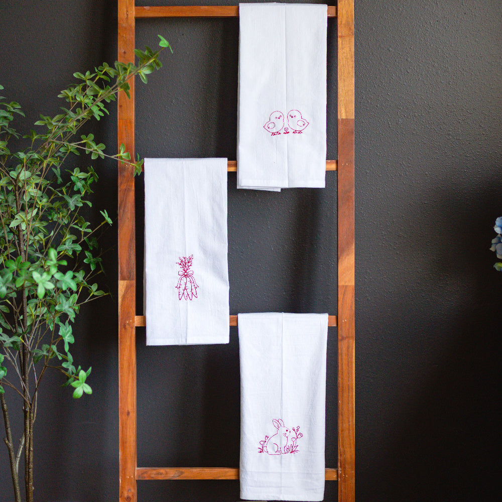 White towels with red embroidery on a wooden ladder against a dark wall with plants and flowers.
