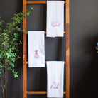 White towels with red embroidery on a wooden ladder against a dark wall with plants and flowers.