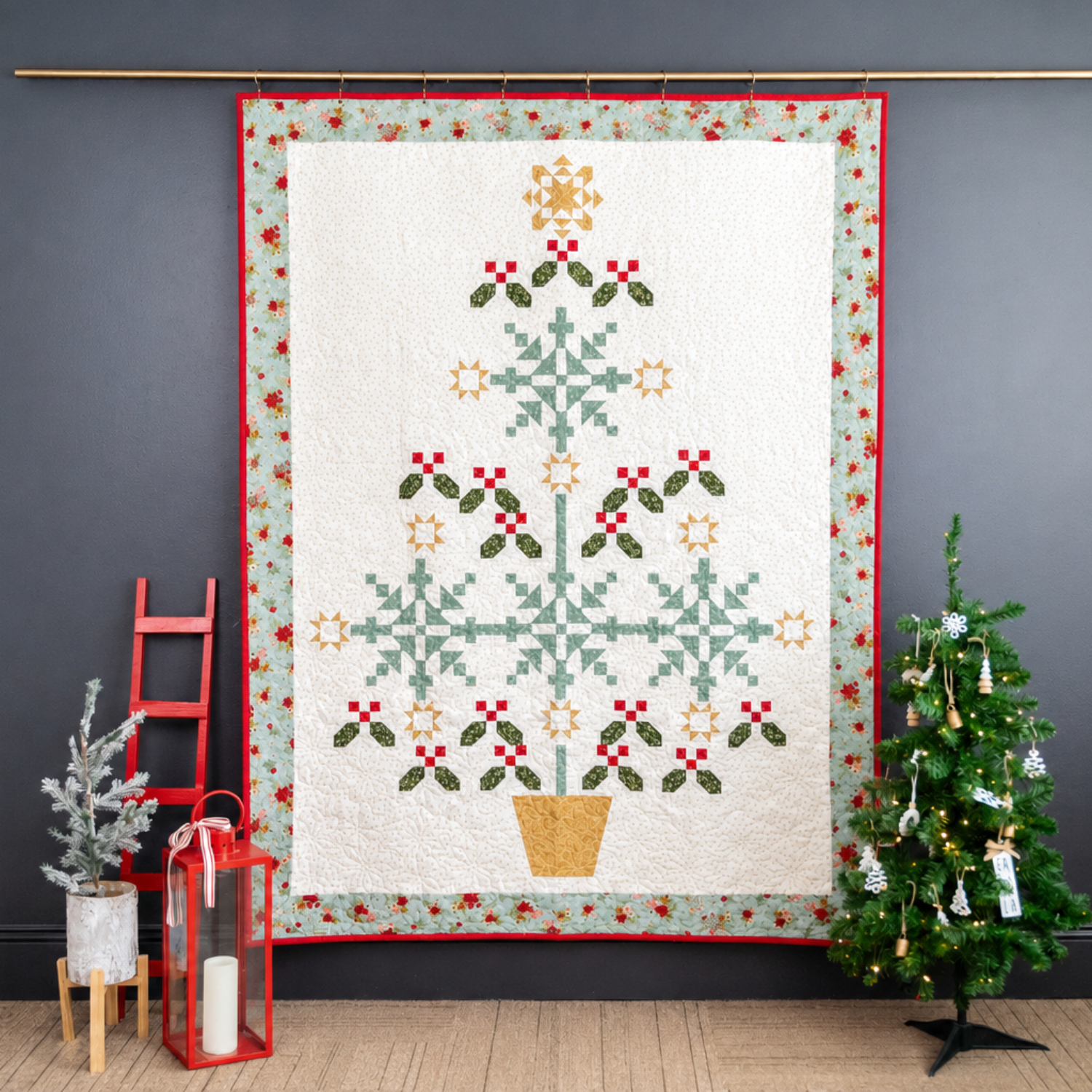 Quilt with Christmas tree design hanging on a wall next to a small decorated tree.