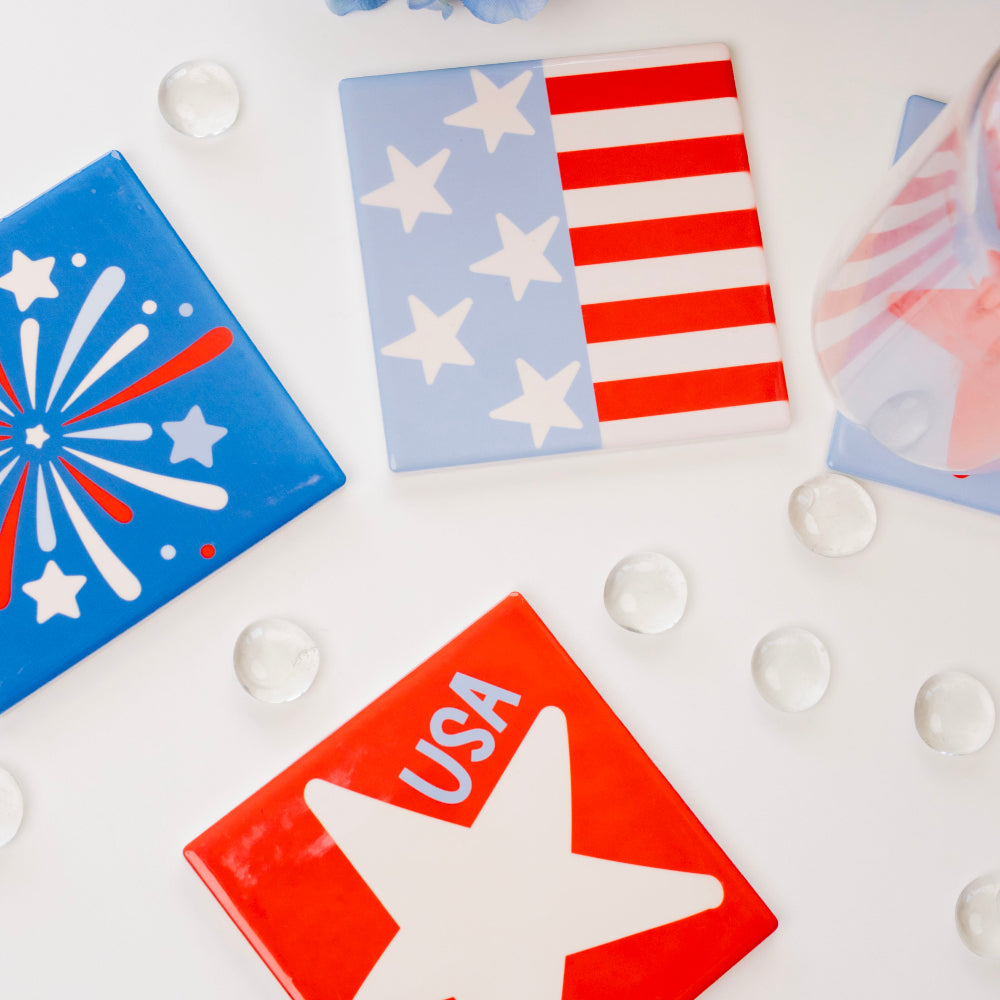 Set of patriotic coasters with American flag, star, and firework designs on a white surface with flowers in the background.