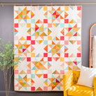 A colorful geometric quilt hanging on a gold rod against a dark gray wall, styled in a room with a yellow armchair, a decorative mirror, and a small potted plant.
