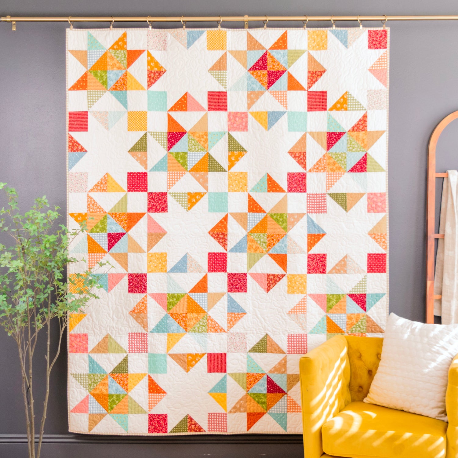 A colorful geometric quilt hanging on a gold rod against a dark gray wall, styled in a room with a yellow armchair, a decorative mirror, and a small potted plant.