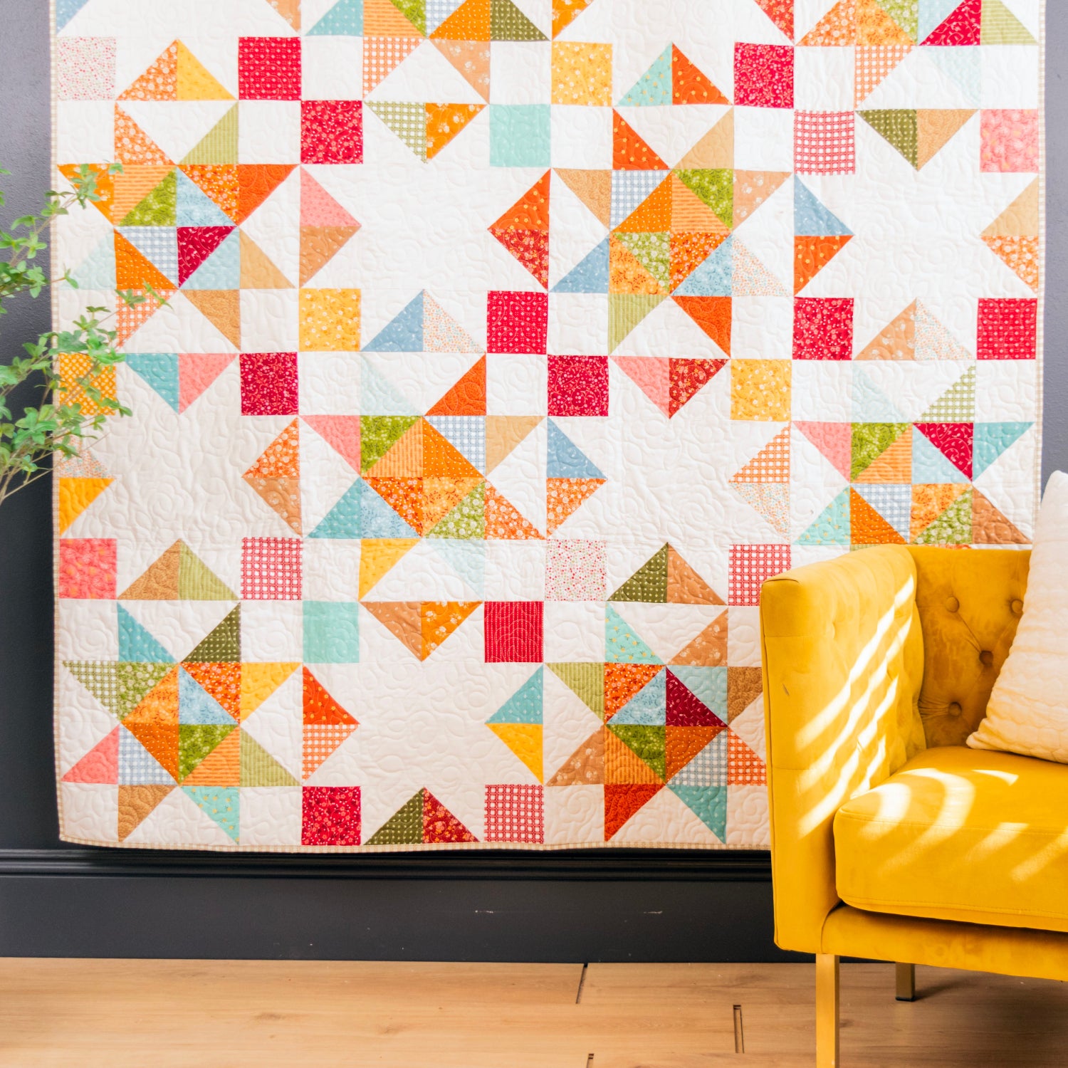 A colorful geometric patchwork quilt hanging on a dark gray wall in a styled room, next to a yellow armchair with a white pillow and a potted plant.