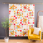 A colorful geometric quilt hanging on a wall in a styled room, with a yellow armchair, potted plant, and wooden ladder shelf nearby.