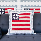 Quilted pillow with American flag design on a gray sofa, with quilted wall art in the background.