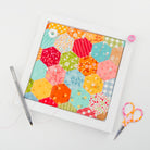 A square white-framed art print or canvas featuring a colorful patchwork of hexagonal fabric shapes in orange, yellow, red, green, blue, and pink, styled on a white background with a gray pen, pink thread, and small scissors.