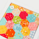 A square framed wall art piece featuring a colorful patchwork of hexagonal fabric panels in floral, striped, and solid patterns, displayed on a clean white background.