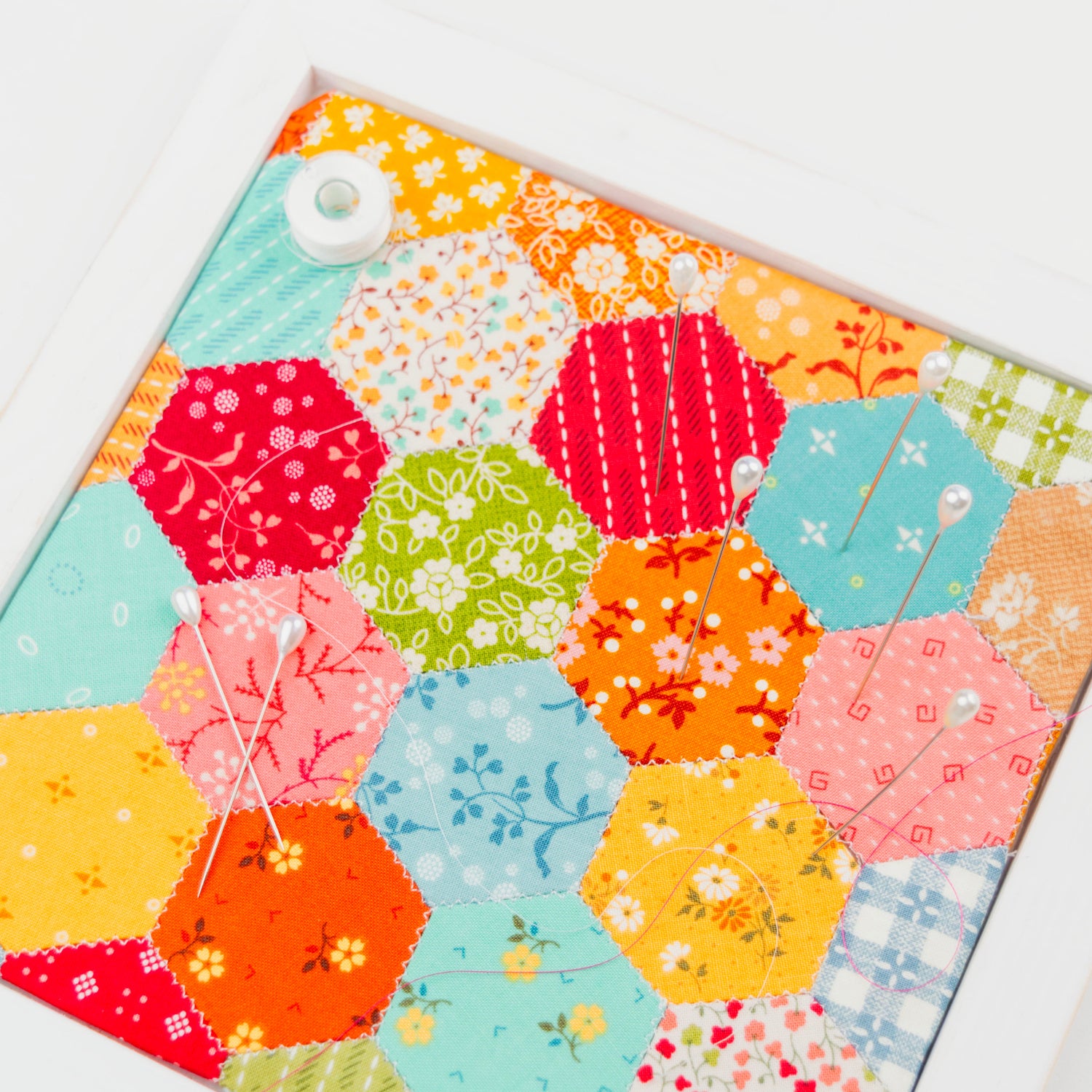 A square framed wall art piece featuring a colorful patchwork of hexagonal fabric panels in floral, striped, and solid patterns, displayed on a clean white background.