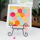 A framed patchwork art print featuring colorful hexagonal quilt blocks, displayed on a black metal easel stand on a white table in a home setting.