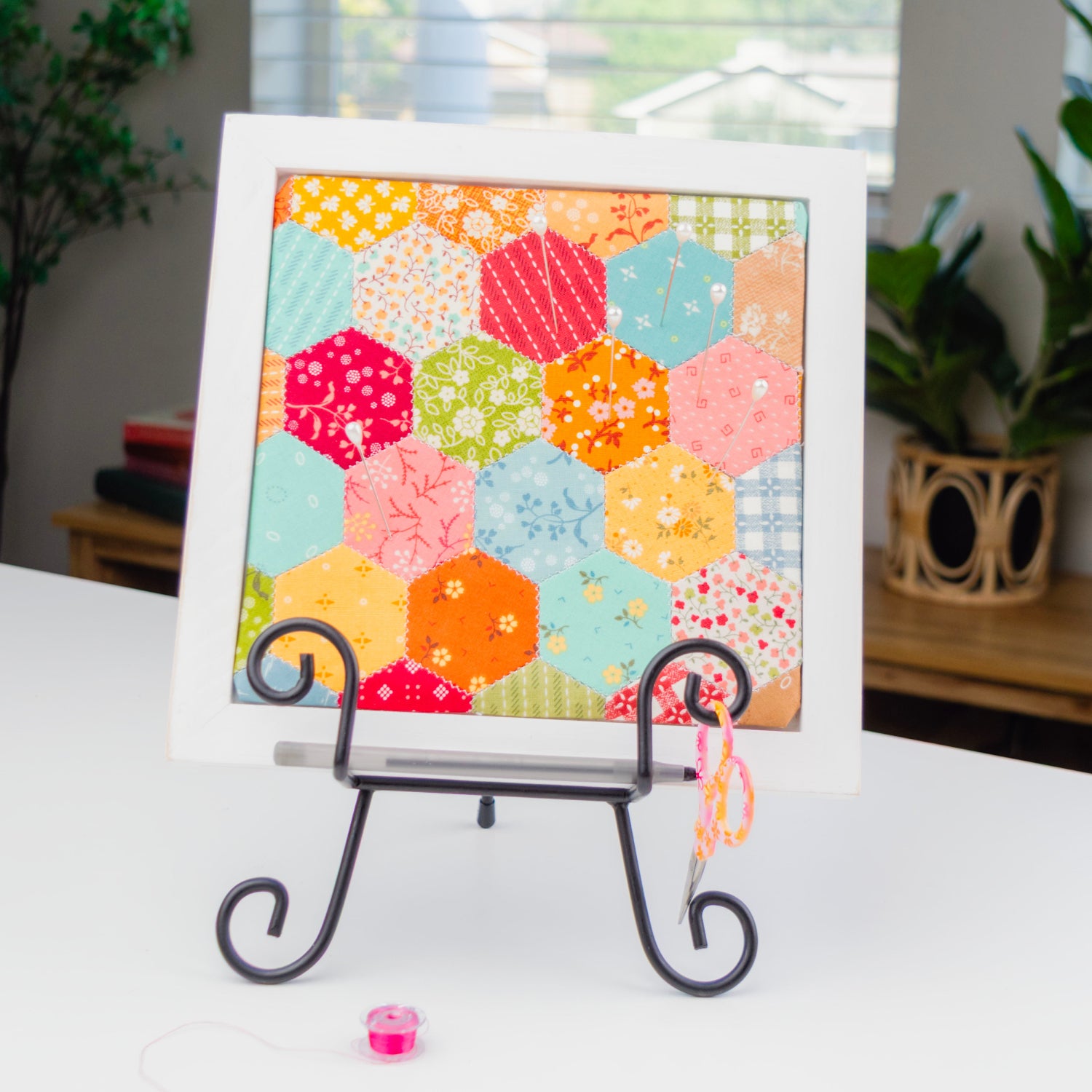 A framed patchwork art print featuring colorful hexagonal quilt blocks, displayed on a black metal easel stand on a white table in a home setting.