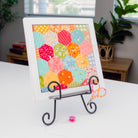 A framed art print featuring a colorful pattern of hexagonal quilt blocks in orange, pink, yellow, green, and blue, displayed on a black metal easel stand on a white table.