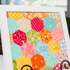 A white-framed hexagonal patchwork quilt art print displayed on a black metal stand in a home setting with a blurred background of plants and books.
