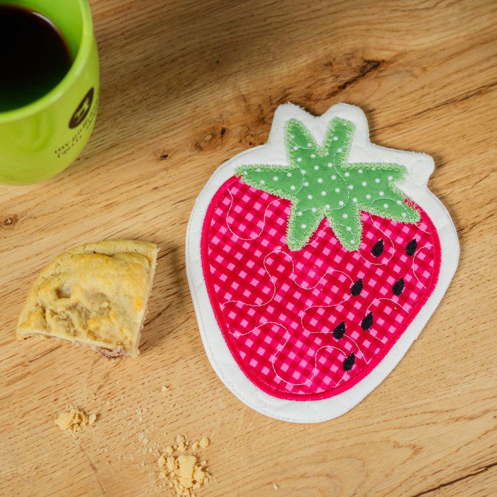 Strawberry-shaped coaster on a wooden surface with a green mug and pastry.