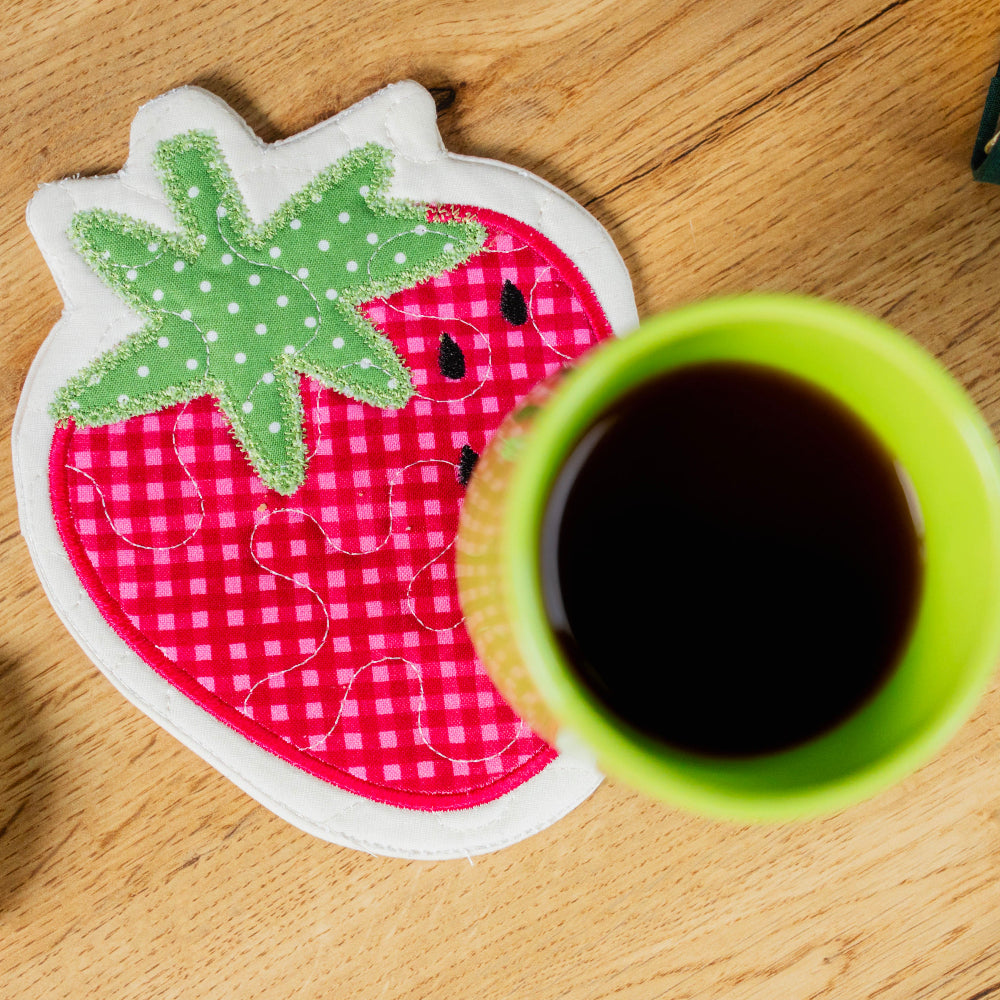 Coaster with strawberry design on a wooden surface next to a cup of coffee.