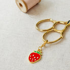 A gold metal key ring with a dangling strawberry charm, placed on a light beige linen surface next to a spool of pink thread.