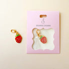 A strawberry-shaped enamel charm with red body, green leaves, and gold-tone clasp, shown next to its pink display card labeled ENAMEL CHARM on a light beige background.