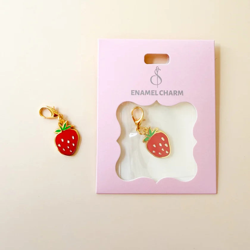 A strawberry-shaped enamel charm with red body, green leaves, and gold-tone clasp, shown next to its pink display card labeled ENAMEL CHARM on a light beige background.