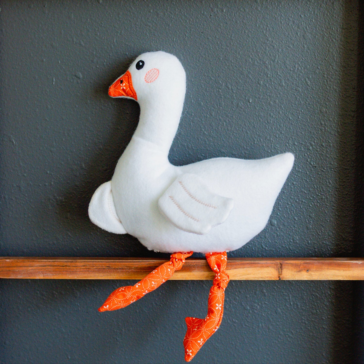 White goose figurine with orange feet and beak on a wooden perch against a gray wall.