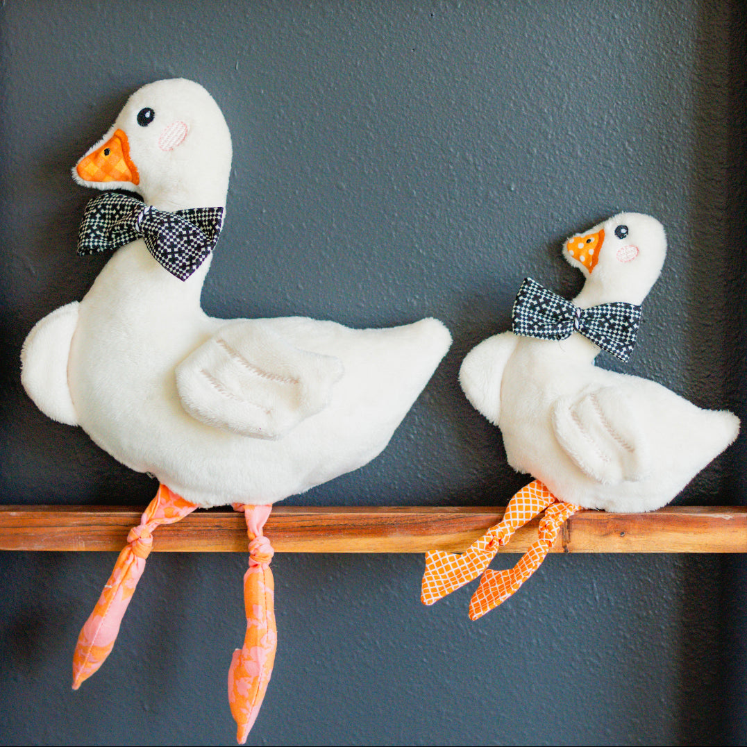 Two decorative goose figures with bow ties on a wooden shelf against a dark wall.