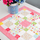 Quilted table runner with colorful patterns on a white table, accompanied by a vase of flowers.