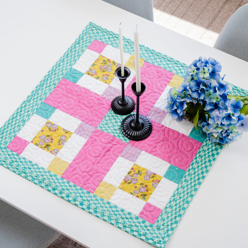 Colorful quilted table runner on a white table with candles and flowers.