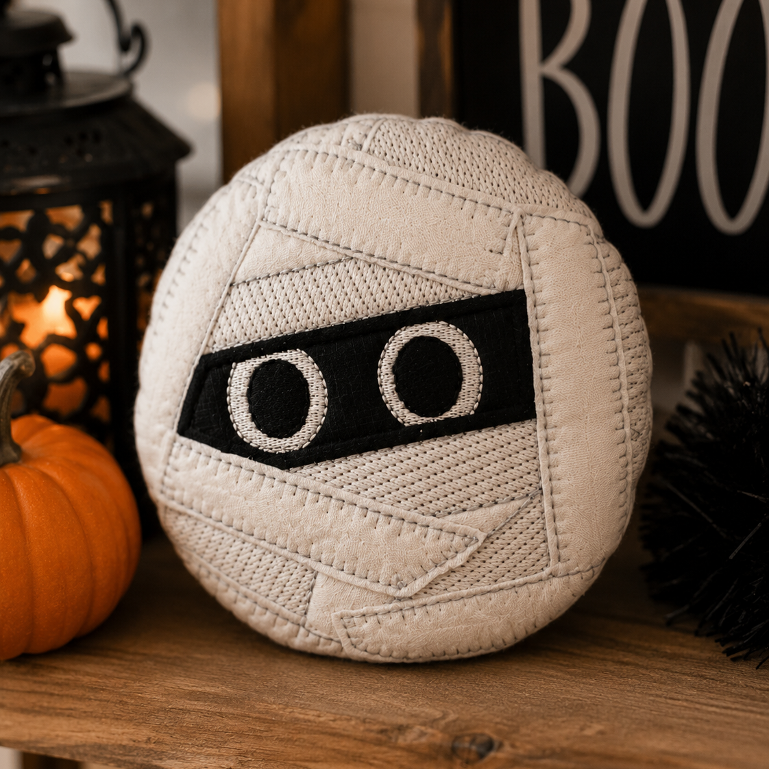 A round mummy-themed decorative pillow with a cream-colored textured wrap design and a black eye patch, displayed on a wooden shelf next to a small orange pumpkin and a black lantern.