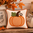 A square cream fabric pillow with an embroidered orange pumpkin design, styled on a wooden surface with autumn decorations including a 'Hello Fall' sign, a knitted pumpkin, and fall leaves.