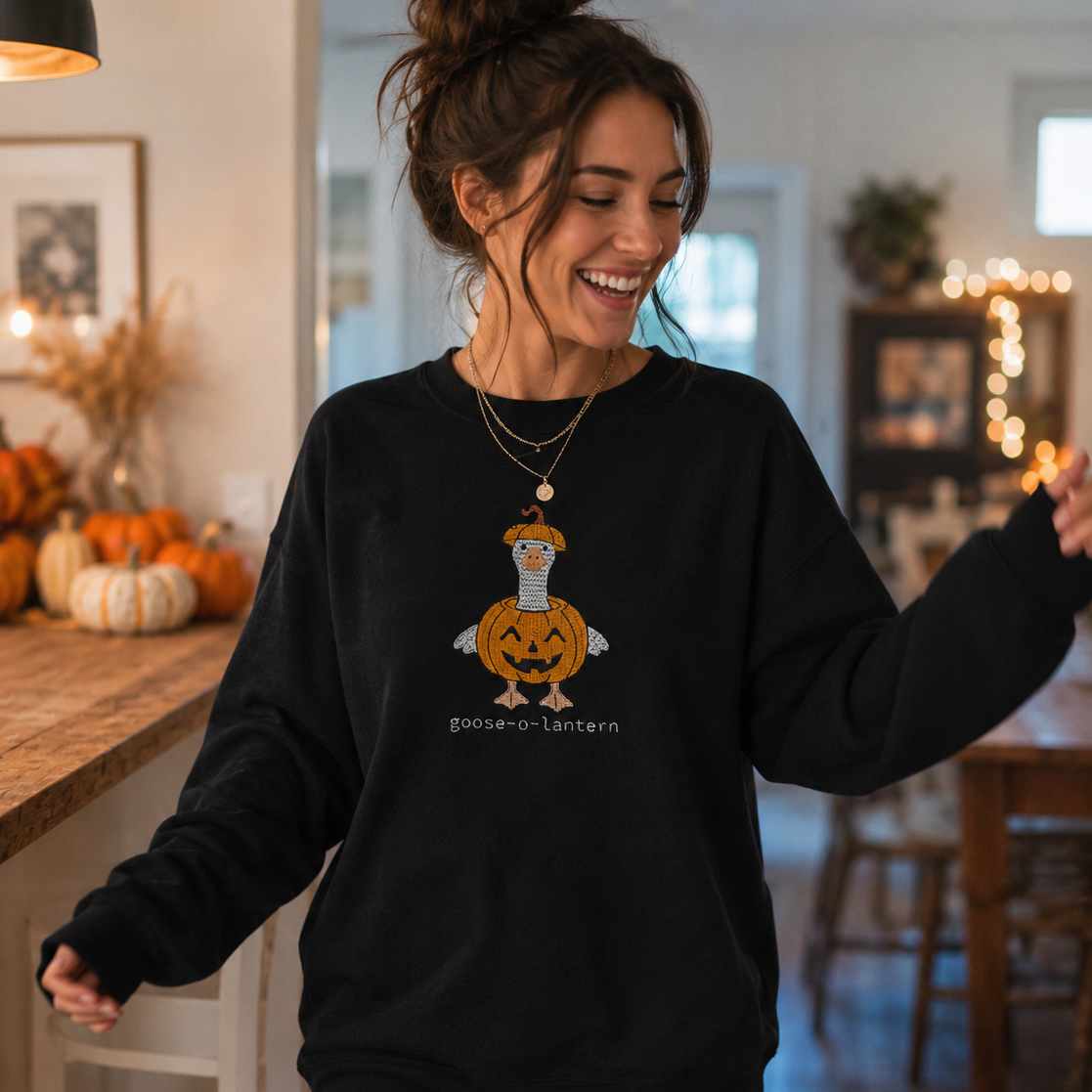 A woman wearing a black crewneck sweatshirt with a cartoon goose wearing a pumpkin lantern graphic and the text 'goose-lantern' printed below it. She is standing in a cozy, warmly lit home interior with autumn decorations.