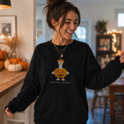 A woman wearing a black crewneck sweatshirt with a cartoon goose wearing a pumpkin lantern graphic and the text 'goose-lantern' printed below it. She is standing in a cozy, warmly lit home interior with autumn decorations.
