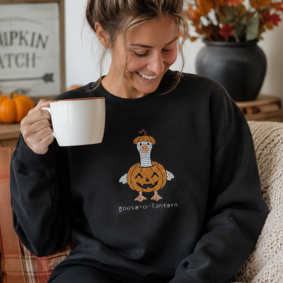 A woman wearing a black crewneck sweatshirt with a graphic of a pumpkin-lantern goose and the text 'Goose-o-Lantern' sits on a couch holding a white mug in a cozy, autumn-themed room.