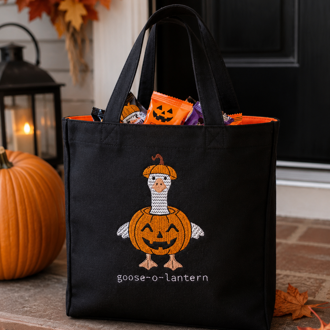 A black fabric Halloween treat bag with a stitched 'goose-o-lantern' design featuring a pumpkin-faced goose and candy peeking out, set on a brick porch next to a pumpkin and autumn leaves.