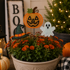 Halloween-themed floral garden stake decorations featuring a jack-o'-lantern, a witch hat, and a ghost, displayed in a pot of orange mums with decorative pumpkins and a lit Christmas tree in the background.