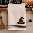 An off-white cotton kitchen towel with a black witch hat and orange band embroidery, plus a black-and-white plaid stripe border at the bottom, hanging on a dresser in a fall-themed setting.