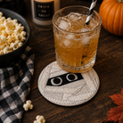 A Halloween-themed tabletop scene with a clear glass tumbler filled with an amber drink and ice, set on a round embroidered coaster. The coaster has a stitched jack-o'-lantern face.