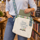 A woman in a bookstore wearing a cream canvas crossbody tote bag with a blue floral strap and 'Keep Turning Pages' embroidery, holding a green book.
