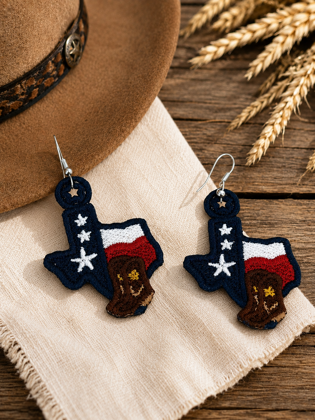 Texas-shaped earrings with American flag design on a wooden surface with a hat and wheat.