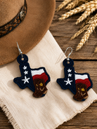 Texas-shaped earrings with American flag design on a wooden surface with a hat and wheat.