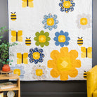 A colorful quilted wall hanging or small quilt displayed in a styled room corner, featuring large yellow flowers, blue and green flower motifs, and bumblebee appliqués on a white background. The room includes a dark gray wall, a yellow armchair, a wooden side table with books and a plant, and a peach-framed chair.