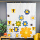 A white quilted wall hanging or blanket featuring colorful floral and bee applique motifs, displayed hanging from a brass rod against a dark gray wall in a styled room with a plant and chair visible.