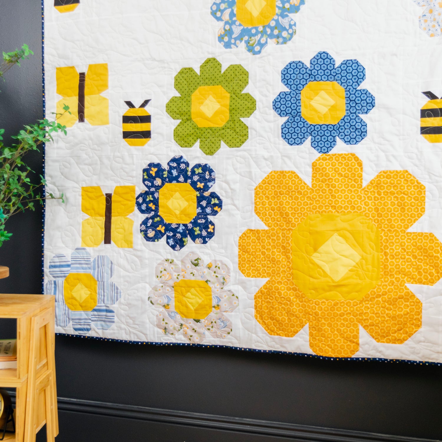 A colorful quilted wall hanging or blanket featuring large yellow and blue flowers, smaller green and blue flowers, bumblebees, and butterflies on a white background, styled in a room next to a wooden side table with a plant.