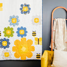 A white quilted wall hanging or blanket with large yellow and blue flower motifs, green flower accents, and small bumblebee designs, displayed in a styled room next to a yellow chair and a wooden floor.