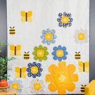 A quilted wall hanging or blanket featuring a whimsical bee and flower pattern in yellow, blue, and green on a white background, styled in a bedroom setting next to a yellow chair and a plant.