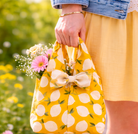 Person holding a yellow reusable bag with lemon pattern filled with flowers, outdoors.