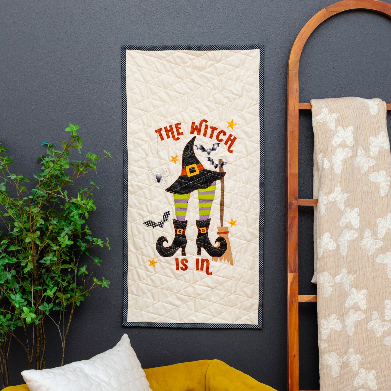 A vertical quilted wall hanging with a Halloween design featuring a witch's hat, broomstick, and the text 'THE WITCH IS IN' is displayed on a dark gray wall next to a wooden ladder shelf holding another quilt.
