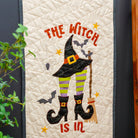 A vertical quilted wall hanging or tapestry with a Halloween witch design, shown hanging on a dark wall next to a wooden ladder holding another quilt. The main design features the words 'THE WITCH IS IN' in orange, a black witch hat, green-and-yellow striped legs, black boots, bats, and stars.