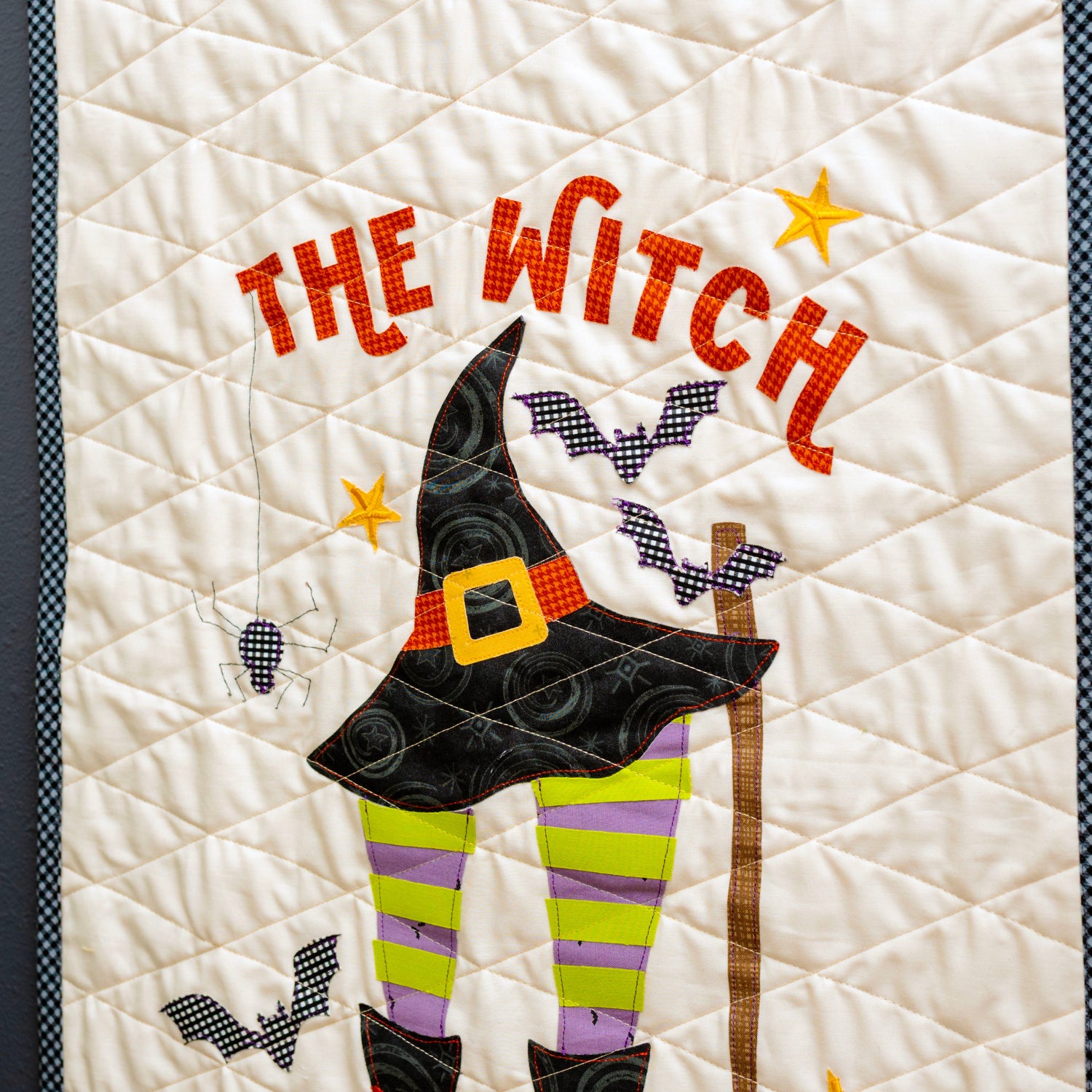 A close-up of a quilted wall hanging or textile panel featuring the words “THE WITCH” in orange, a black witch hat, purple and green striped legs, bats, stars, and a brown staff on an off-white background, photographed against a dark gray wall.